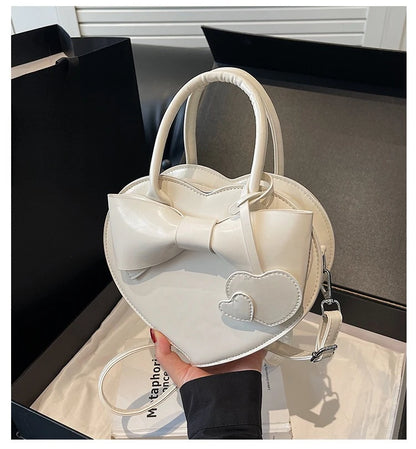 carauana-lovely-shoulder-bag-heart White heart-shaped handbag with a bow detail held by a person.